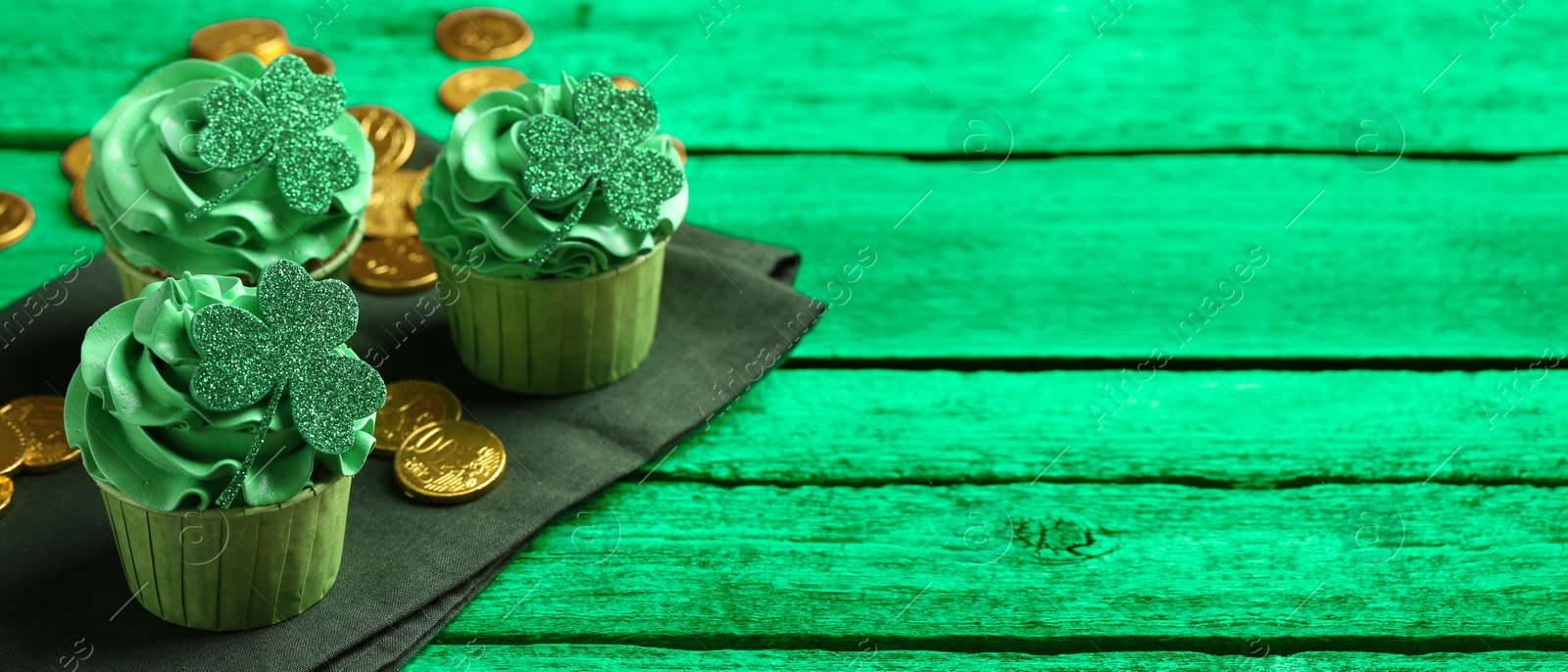 Image of St. Patrick's day. Tasty cupcakes with clover leaf toppers and cream on green wooden table, closeup. Banner design with space for text