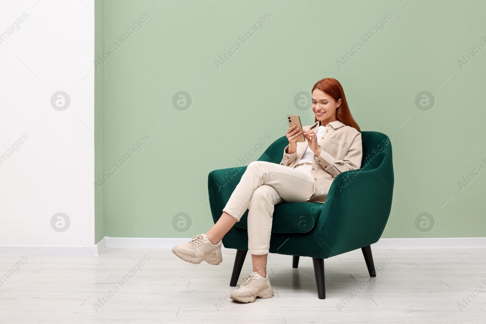 Photo of Happy young woman with smartphone sitting in armchair near light olive wall indoors. Space for text