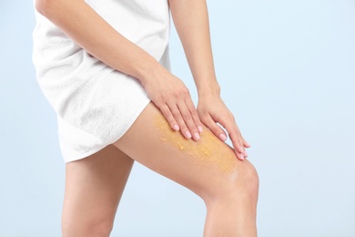 Young woman applying body scrub on leg against light background