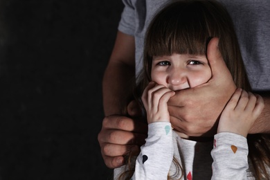 Adult man covering scared little girl's mouth on dark background, space for text. Child in danger