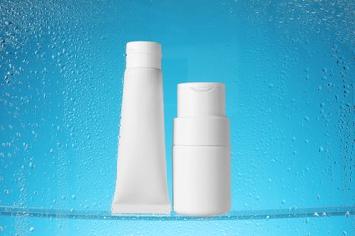Photo of Tube and bottle with moisturizing cream on light blue background, view through wet glass