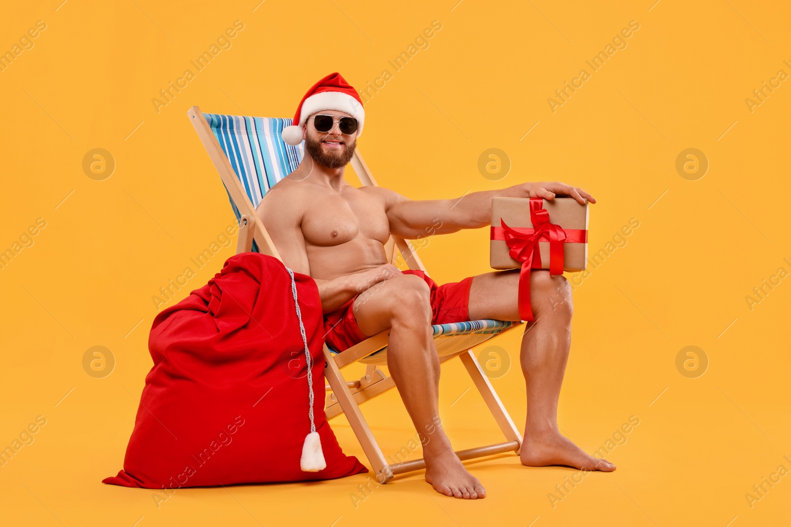 Photo of Muscular young man in Santa hat with deck chair, bag, sunglasses and Christmas gift box on orange background