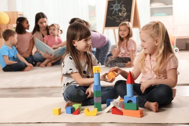 Photo of Cute girls playing with building blocks on floor while kindergarten teacher reading book to other children indoors