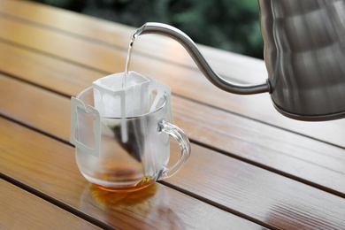 Photo of Pouring hot water into glass cup with drip coffee bag from kettle on wooden table, closeup