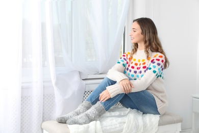 Beautiful teenage girl in warm cozy sweater looking out window at home. Space for text