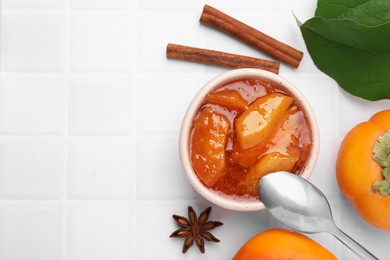 Photo of Bowl of tasty persimmon jam and ingredients on white tiled table, flat lay. Space for text