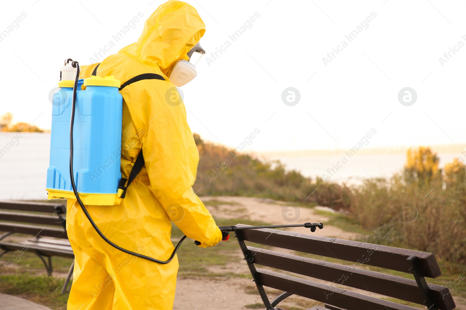 Photo of Person in hazmat suit disinfecting bench in park with sprayer, back view. Surface treatment during coronavirus pandemic