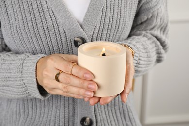 Photo of Woman  with stylish jewelry holding burning soy candle in ceramic holder indoors, closeup