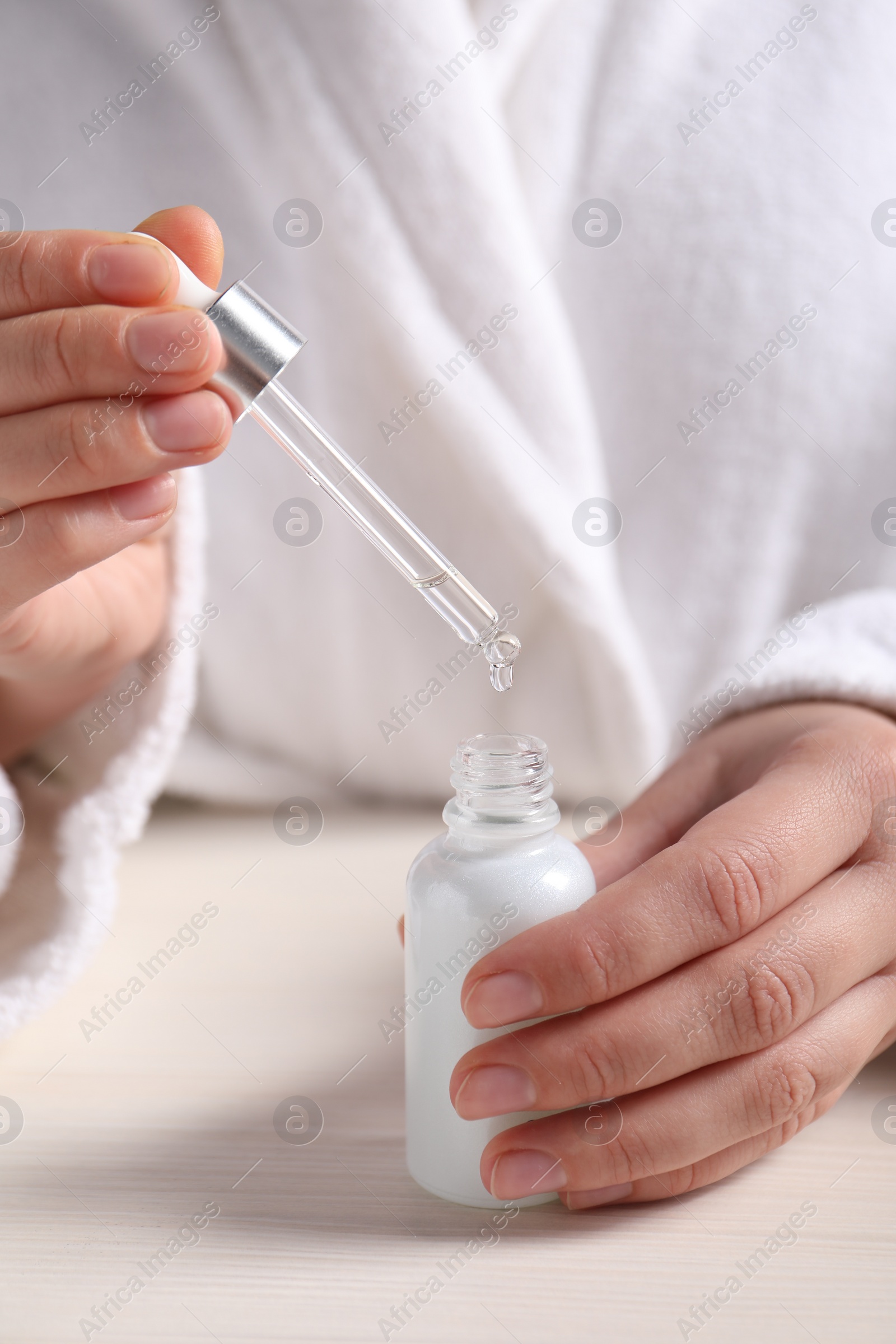Photo of Woman dripping cosmetic serum from pipette into bottle at white table, closeup