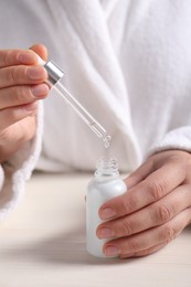 Photo of Woman dripping cosmetic serum from pipette into bottle at white table, closeup
