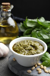 Photo of Tasty pesto sauce in bowl, basil, pine nuts, garlic and oil on grey table
