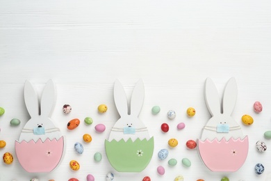 Photo of Candies and bunny figures in protective masks on white wooden table, flat lay with space for text. Easter holiday during COVID-19 quarantine