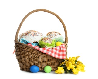 Photo of Basket with delicious Easter cakes, dyed eggs and flowers on white background