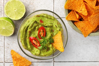 Photo of Bowl of delicious guacamole with chili pepper, nachos chips and lime on white tiled table, flat lay