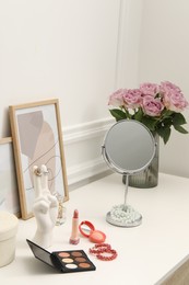 Photo of Mirror, cosmetic products, perfume and vase with pink roses on white dressing table in makeup room