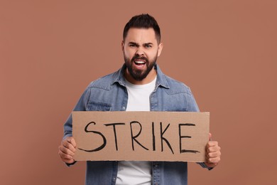 Photo of Angry young man holding cardboard banner with word Strike on brown background