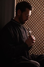 Photo of Catholic priest in cassock holding cross in confessional booth