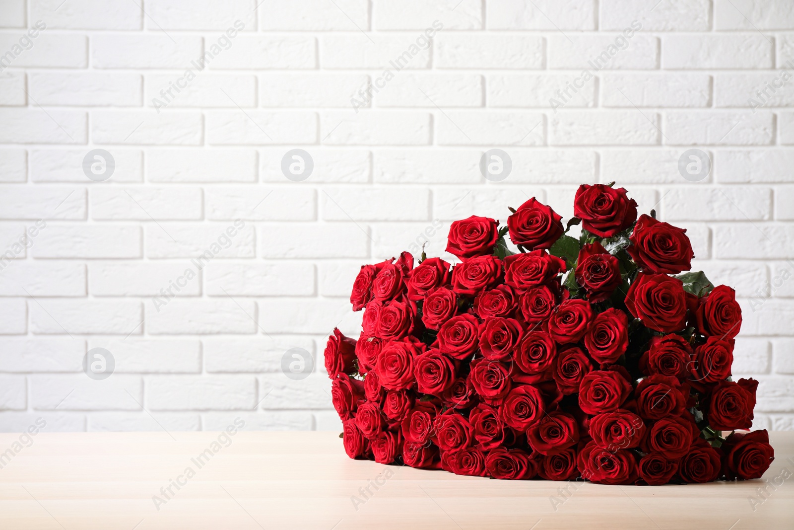 Photo of Luxury bouquet of fresh red roses on wooden table near white brick wall. Space for text