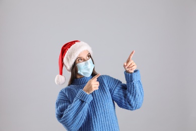 Photo of Pretty woman in Santa hat and medical mask pointing on grey background