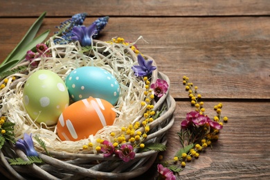 Wicker nest with painted Easter eggs and flowers on wooden table, space for text