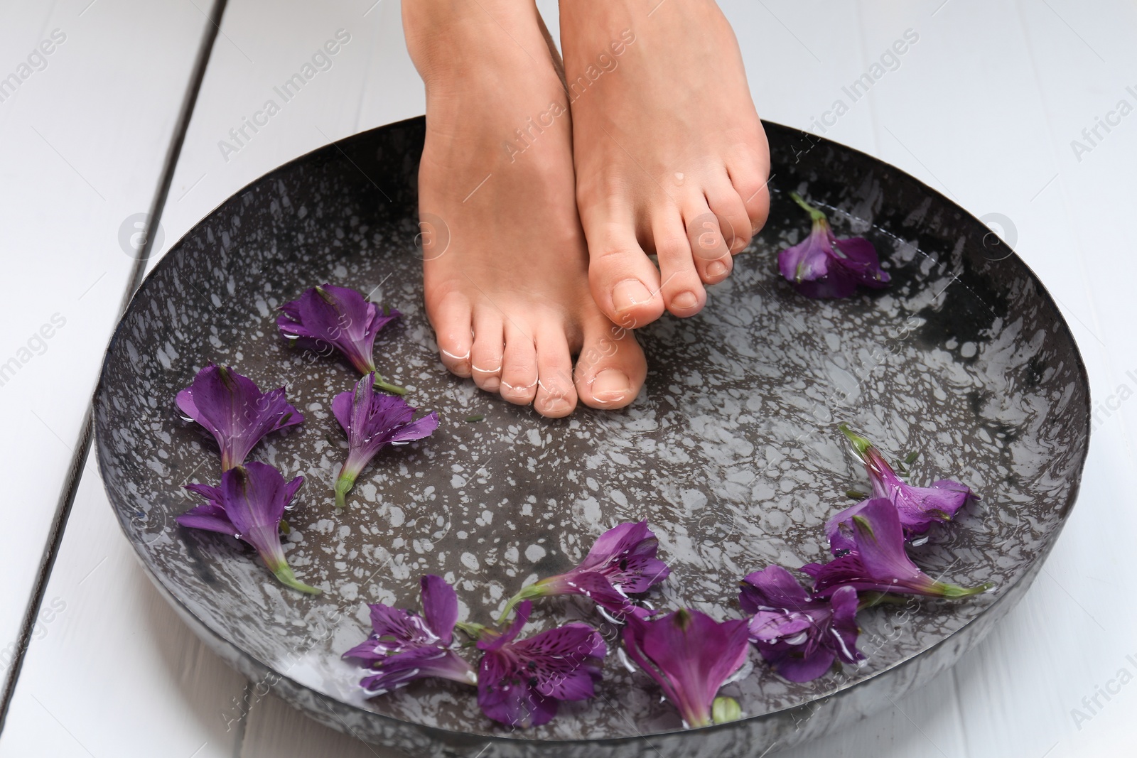 Photo of Woman putting her feet into bowl with water and flowers on white wooden floor, closeup. Spa treatment