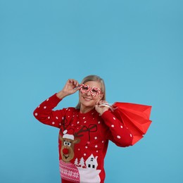 Photo of Senior woman in Christmas sweater and funny glasses with shopping bags on light blue background