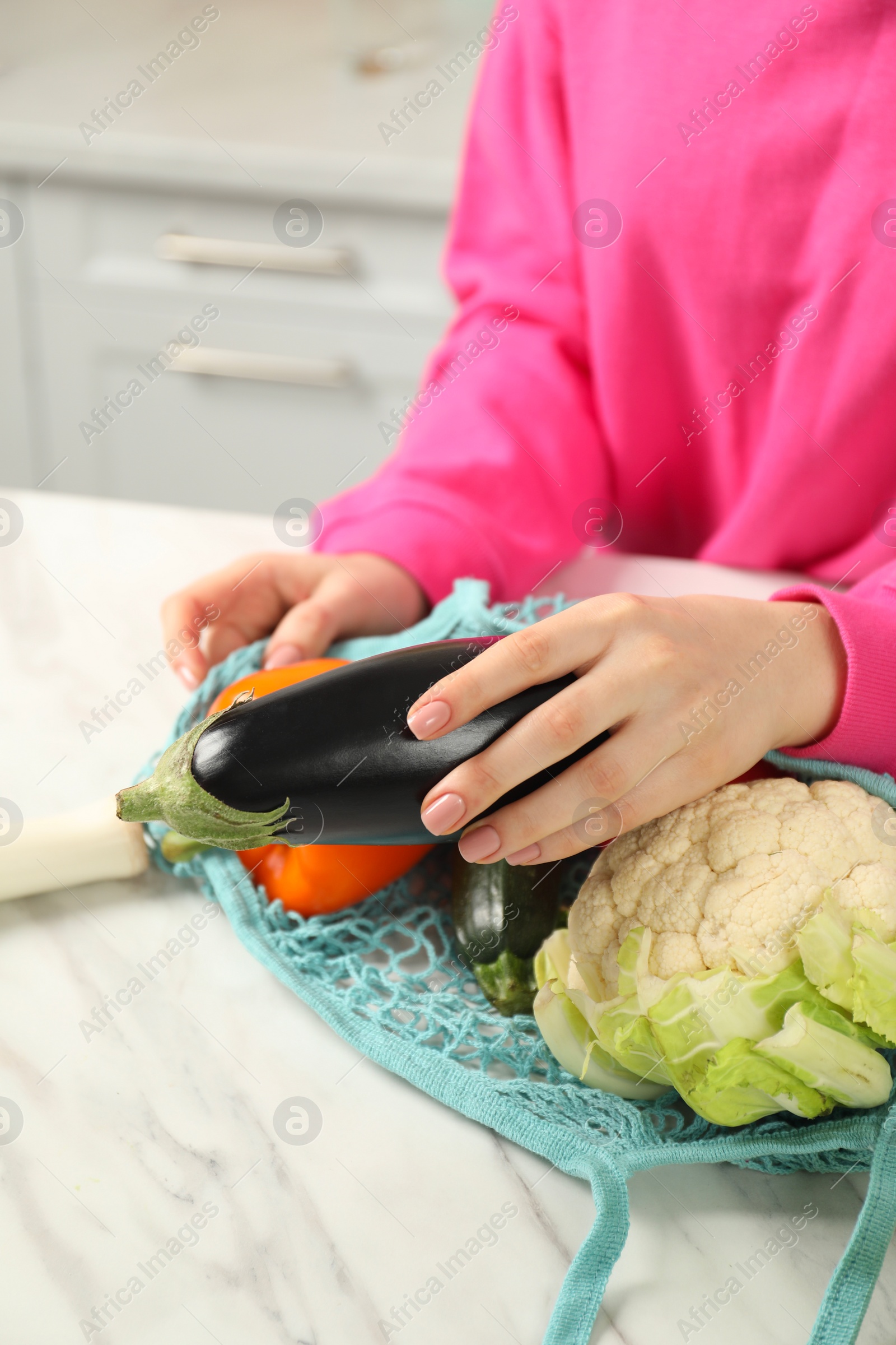 Photo of Woman taking eggplant out from string bag at light marble table, closeup