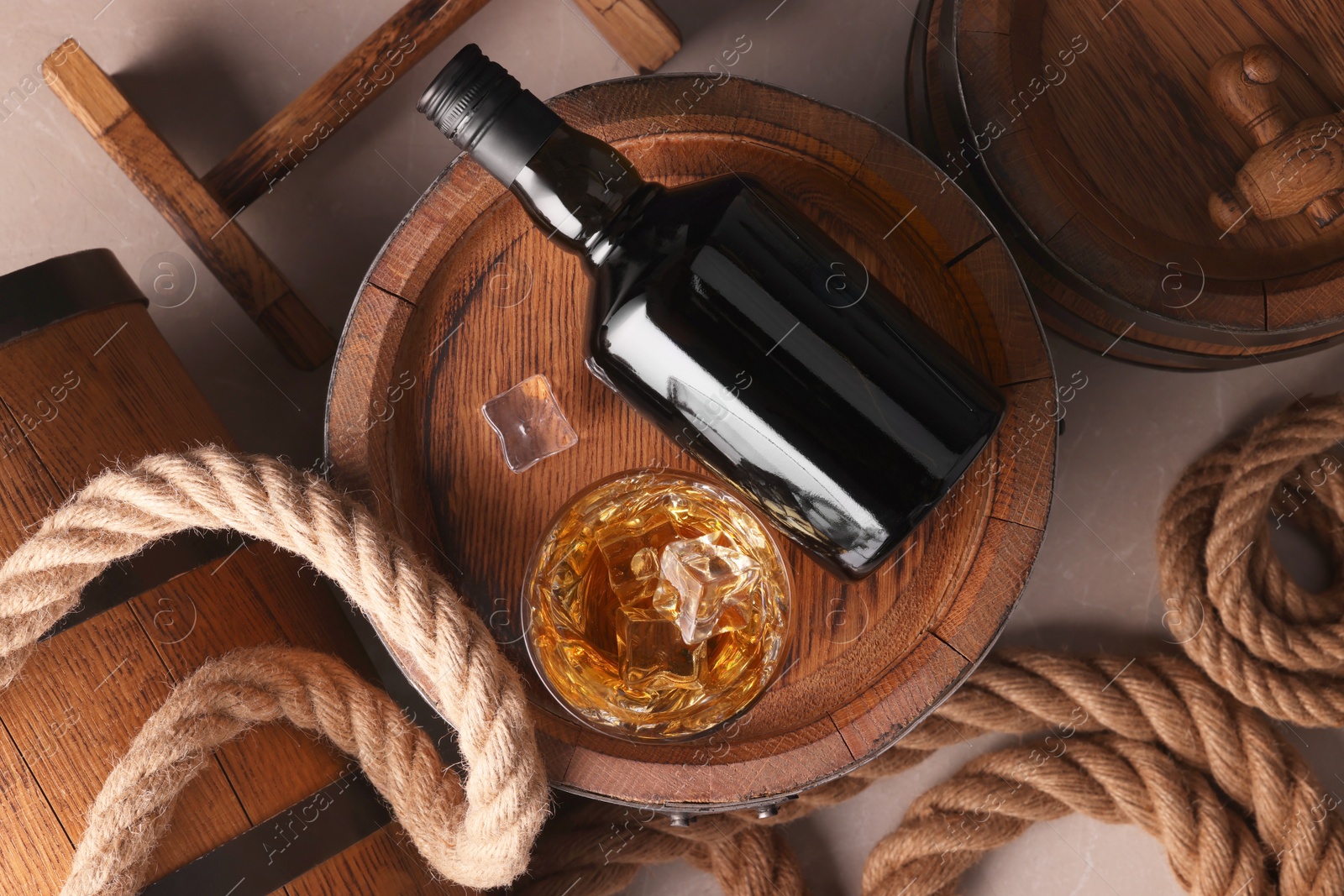 Photo of Whiskey with ice cubes in glass, bottle, wooden barrels and rope on grey marble table, flat lay