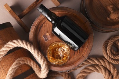 Whiskey with ice cubes in glass, bottle, wooden barrels and rope on grey marble table, flat lay