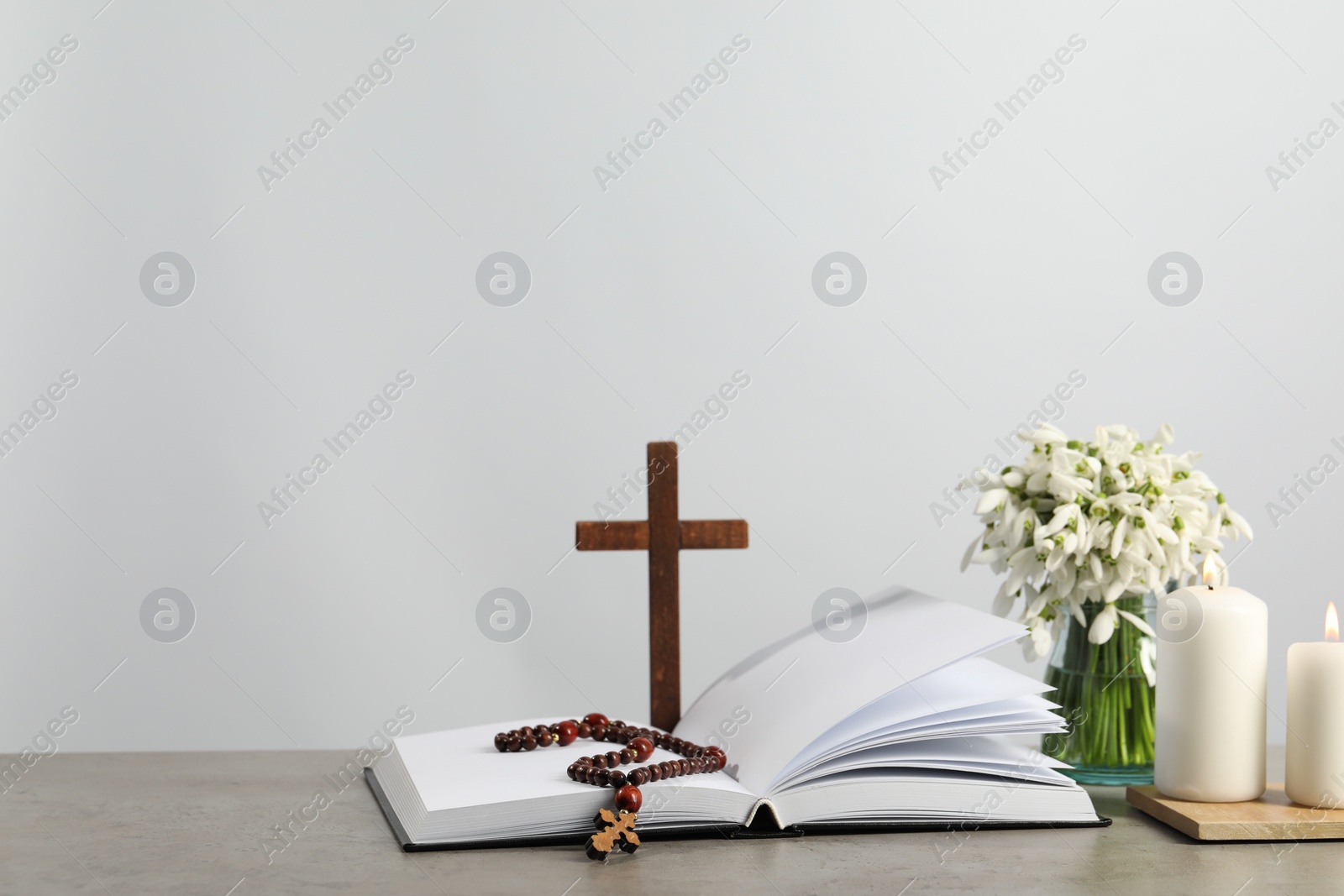 Photo of Church candles, Bible, wooden cross, rosary beads and flowers on grey table. Space for text