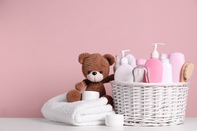 Photo of Baby cosmetic products in wicker basket, bath accessories and knitted toy bear on white table against pink background. Space for text