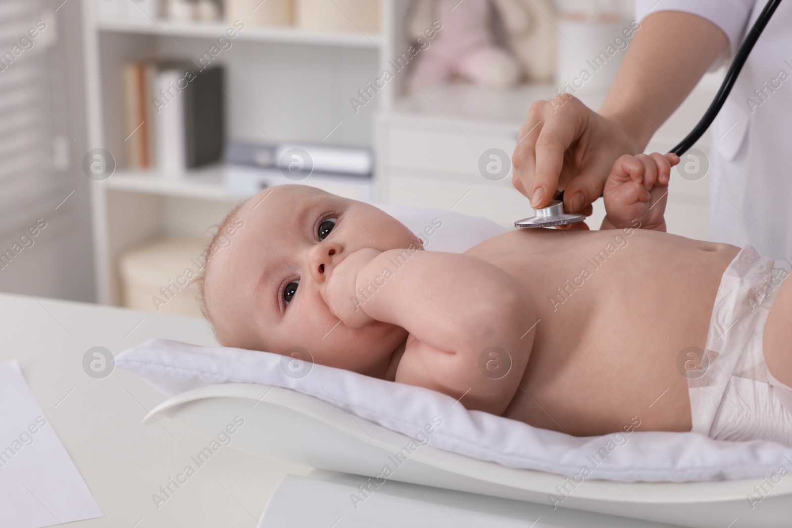 Photo of Pediatrician examining cute little baby with stethoscope and weighting it in clinic, closeup