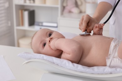 Pediatrician examining cute little baby with stethoscope and weighting it in clinic, closeup