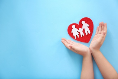 Child protecting red heart with paper silhouette of people on color background, top view