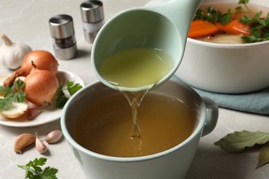 Pouring hot delicious bouillon into cup on light grey table, closeup