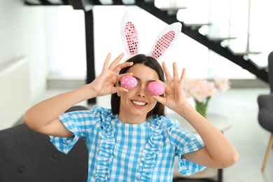 Beautiful woman in bunny ears headband holding Easter eggs near eyes at home