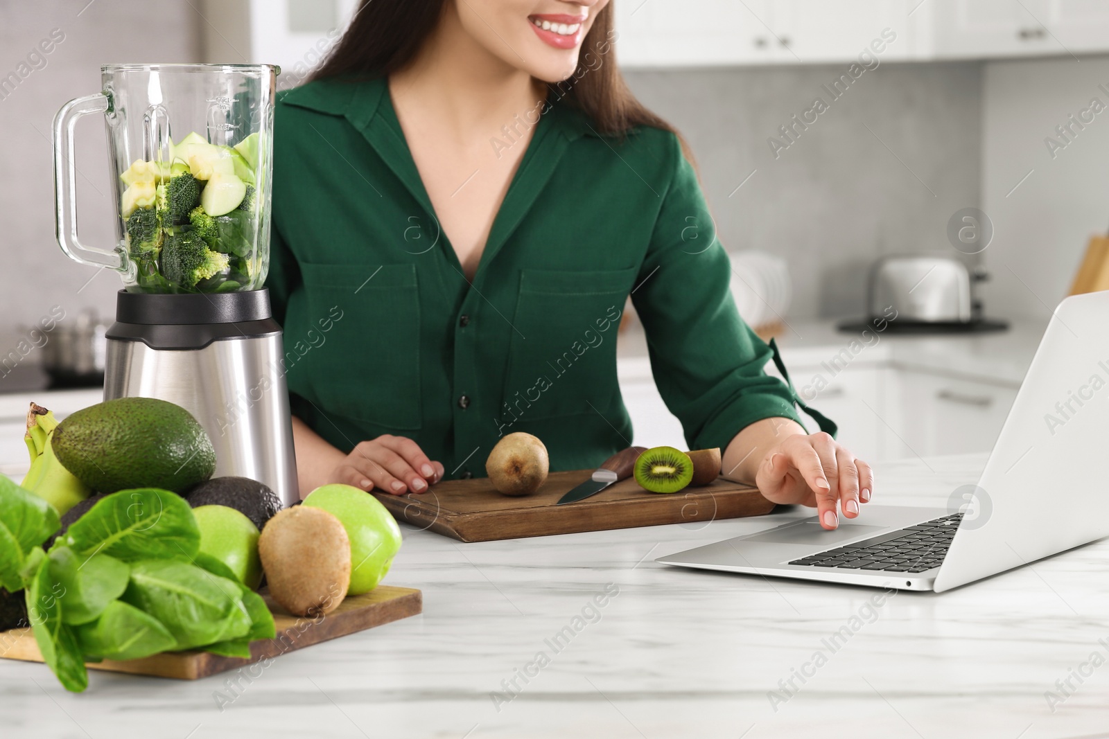 Photo of Young woman using laptop while preparing smoothie at white table in kitchen, closeup