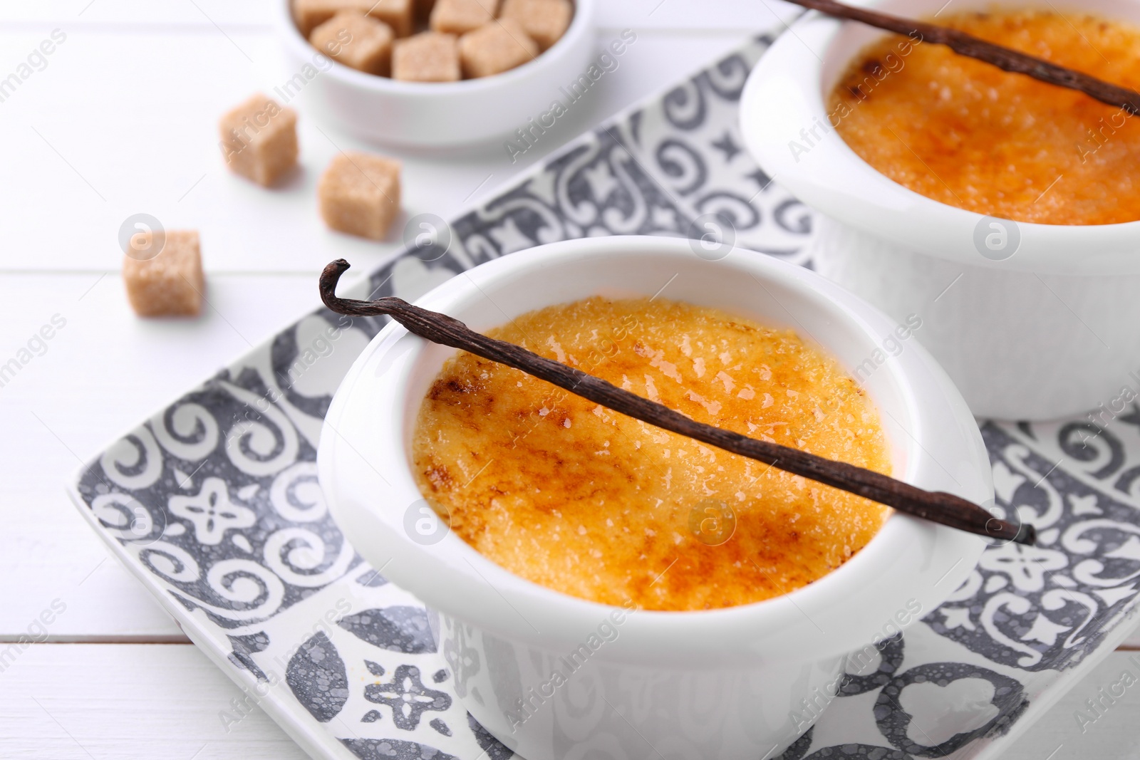 Photo of Delicious creme brulee in bowls, vanilla pods and sugar cubes on white wooden table, closeup