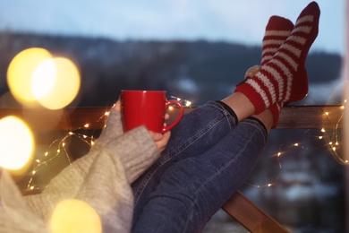 Woman with cup of hot beverage and Christmas lights resting on balcony. Winter evening