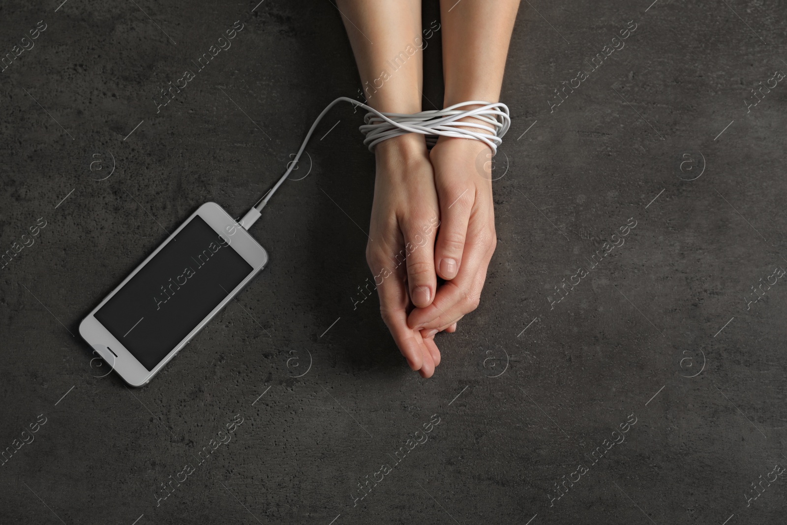 Photo of Woman holding hands tied with charging cable near mobile phone on grey background, above view. Loneliness concept