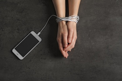 Woman holding hands tied with charging cable near mobile phone on grey background, above view. Loneliness concept