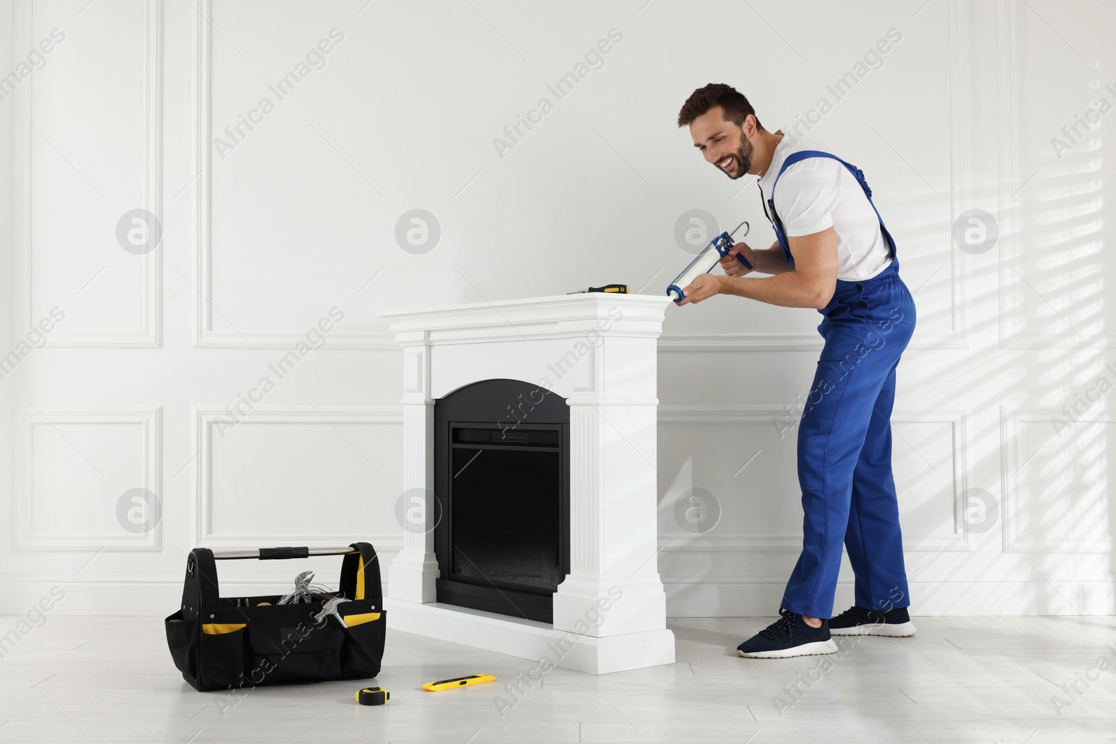 Photo of Professional technician sealing electric fireplace with caulk near white wall in room