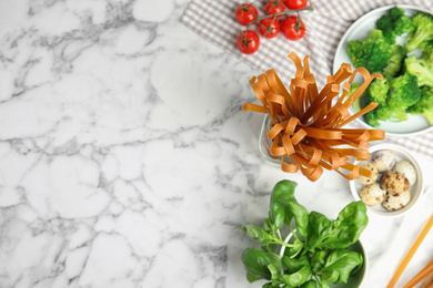 Photo of Uncooked buckwheat noodles and fresh ingredients on white marble table, flat lay. Space for text