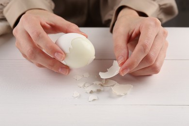 Woman peeling boiled egg at white wooden table, closeup