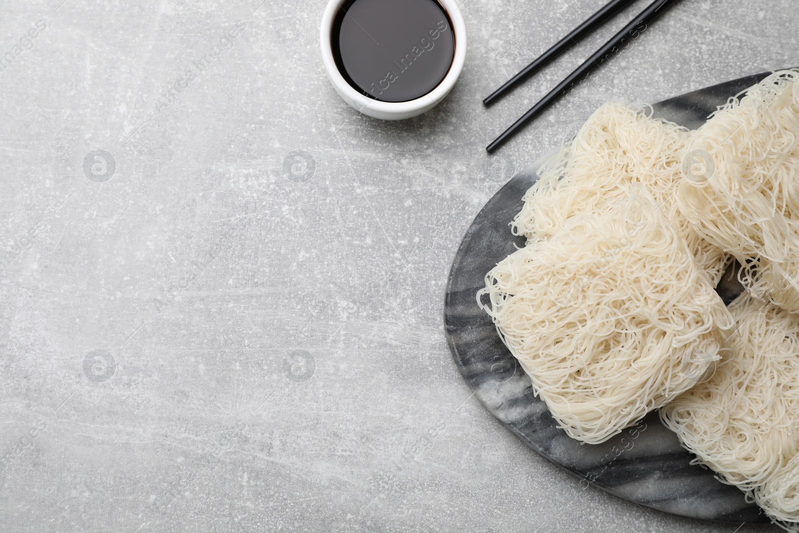 Photo of Uncooked rice noodles, soy sauce and chopsticks on light grey table, flat lay. Space for text