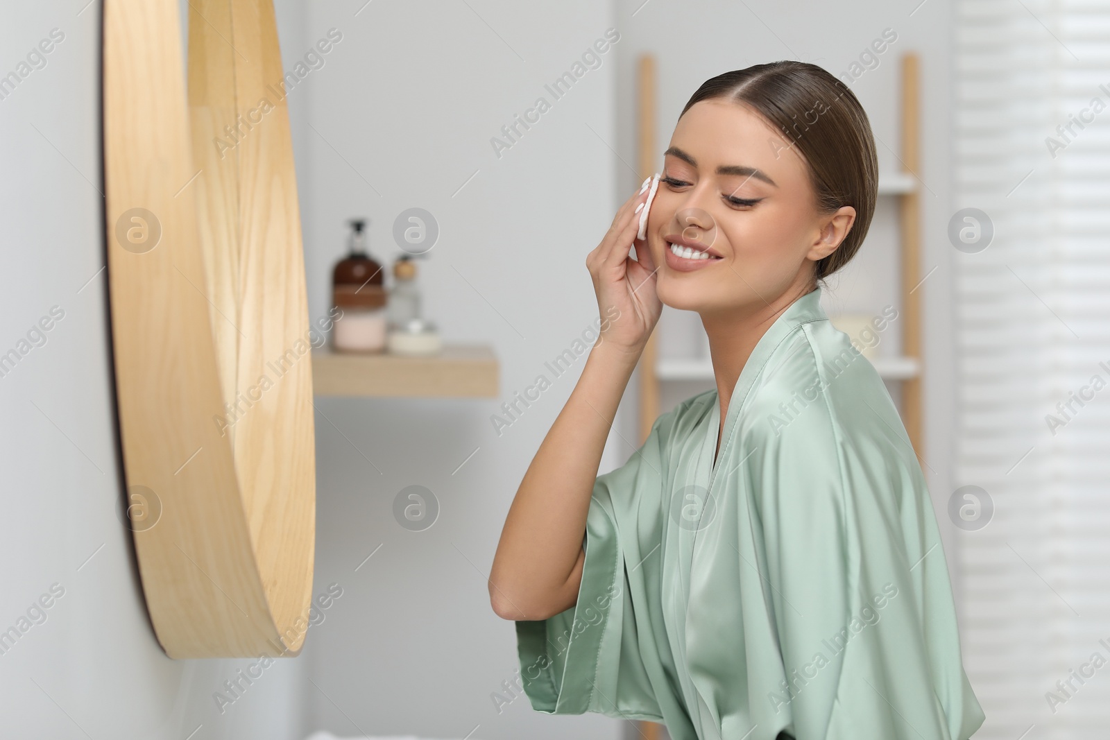 Photo of Beautiful woman removing makeup with cotton pad near mirror indoors