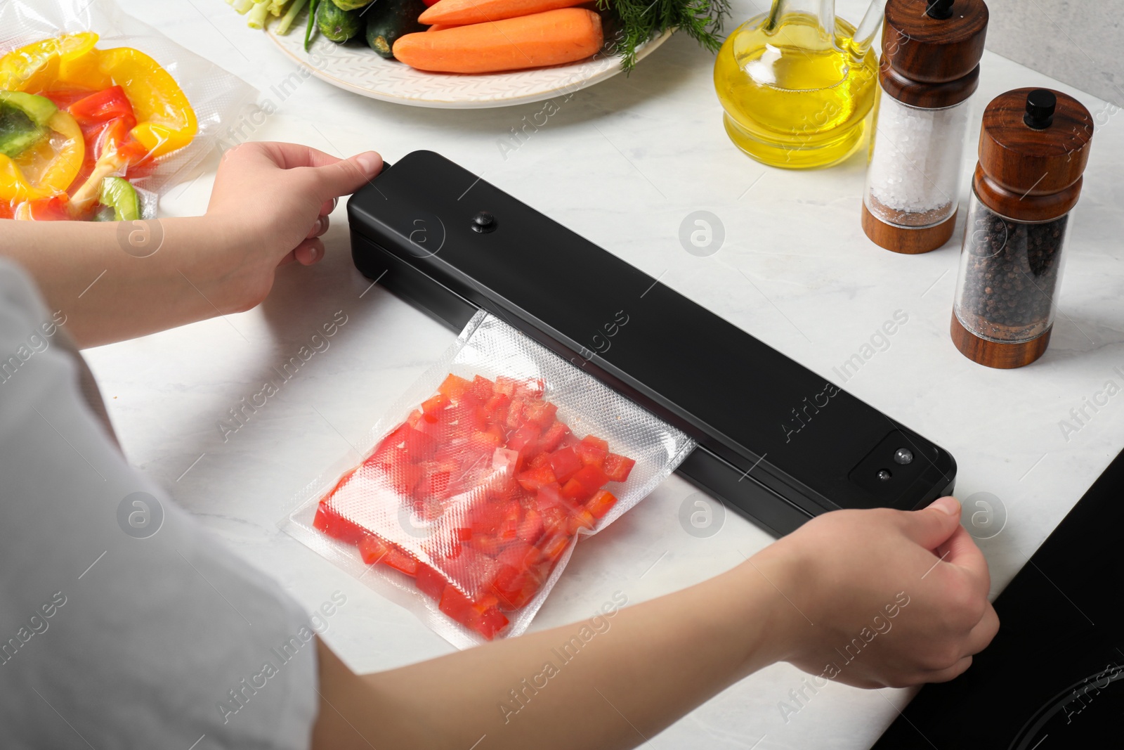 Photo of Woman using sealer for vacuum packing with plastic bag of red pepper at white table, closeup