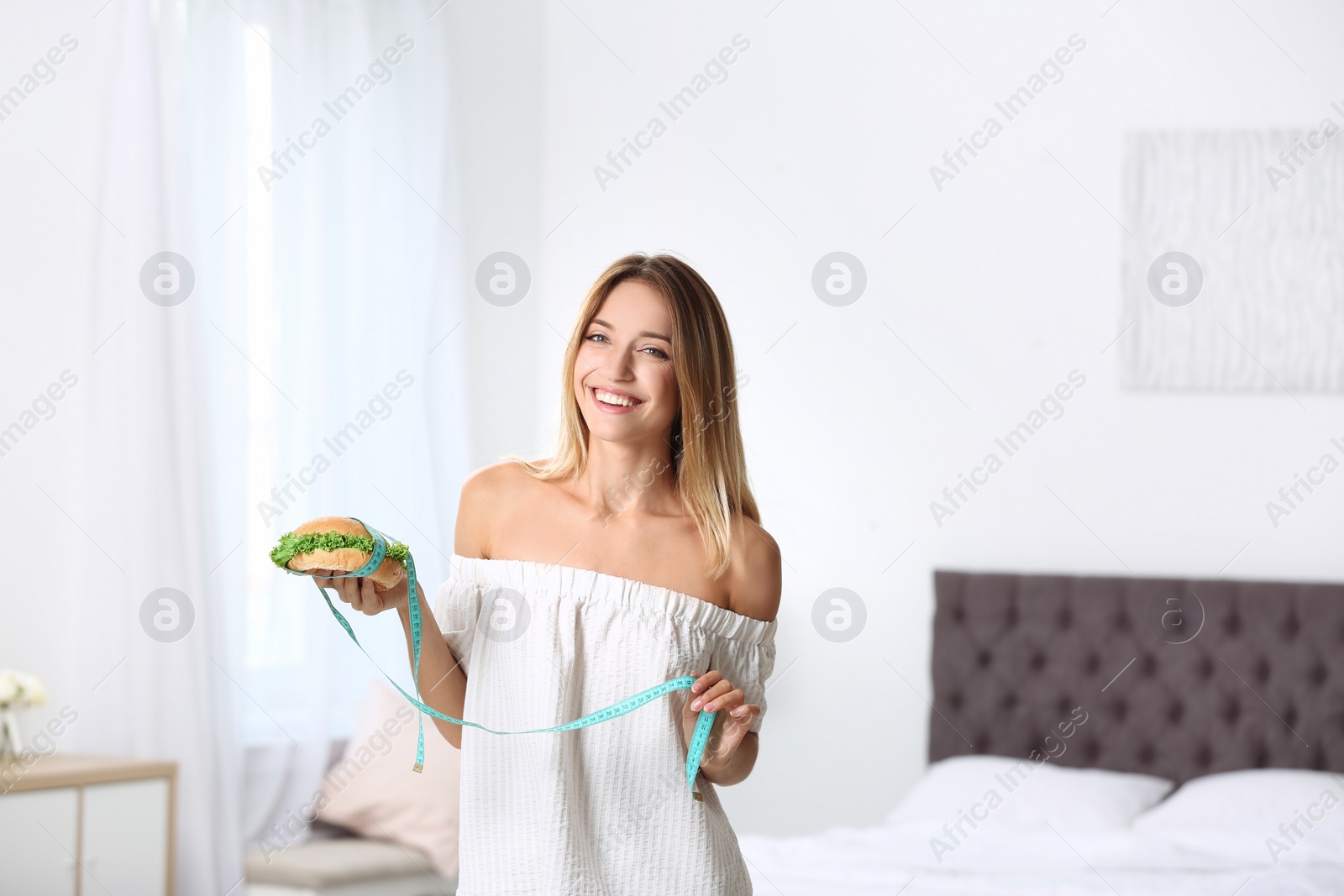 Photo of Happy slim woman with measuring tape and burger at home. Positive weight loss diet results