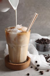 Pouring milk into glass with refreshing iced coffee at gray table, closeup