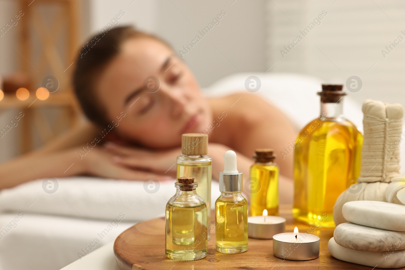 Photo of Aromatherapy. Woman relaxing on massage couch in spa salon, focus on bottles of essential oils, herbal bag, burning candle and stones